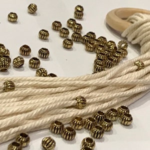 May include: A collection of small, round, metallic gold-toned beads with vertical ribbed textures scattered across a surface alongside a bundle of off-white cotton cord looped through a smooth, circular, light-colored wooden ring.