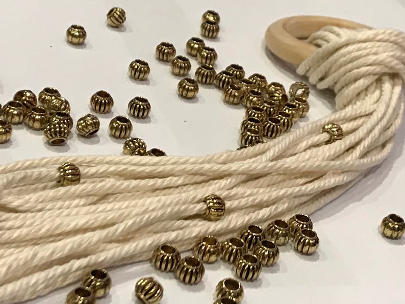 May include: A collection of small, round, metallic gold-toned beads with vertical ribbed textures scattered across a surface alongside a bundle of off-white cotton cord looped through a smooth, circular, light-colored wooden ring.