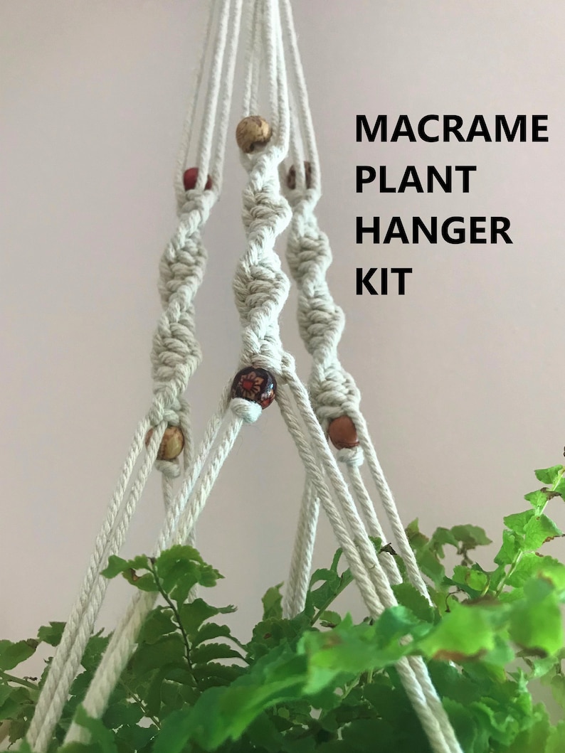 May include: A macrame plant hanger kit featuring off-white cotton cord knotted in a spiral pattern with decorative wooden beads in various brown and red tones. The text MACRAME PLANT HANGER KIT is printed in black on the right side of the image.