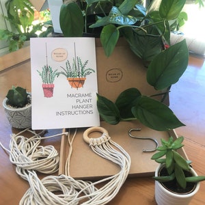 May include: A macram&eacute; plant hanger kit including a white instruction booklet with illustrations and the text Wilde at Home Macram&eacute; Plant Hanger Instructions, a wooden ring with attached cotton cords, a metal S-hook, and two small potted green plants.
