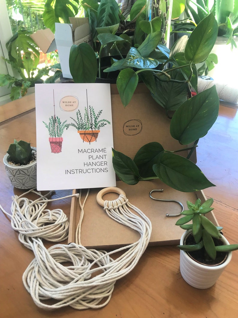 May include: A macram&eacute; plant hanger kit including a white instruction booklet with illustrations and the text Wilde at Home Macram&eacute; Plant Hanger Instructions, a wooden ring with attached cotton cords, a metal S-hook, and two small potted green plants.