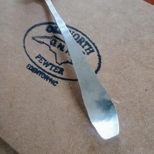 May include: A handcrafted pewter spoon with a hammered texture, resting on a brown surface. The spoon's handle is long and slender, with a rounded bowl. The background features a stamped logo that reads "Old North Pewter, Edenton NC."