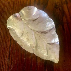 May include: A translucent, leaf-shaped dish with a textured, iridescent surface. The object features a curved, organic form with a pointed tip and a rounded base, exhibiting a pearlescent sheen that reflects light across its uneven, molded glass-like surface.