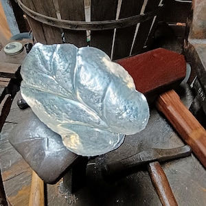 May include: A handcrafted, silver-colored metal leaf-shaped bowl. The bowl sits on a dark metal anvil, surrounded by blacksmithing tools, including hammers and a wooden basket in the background. The bowl's surface has a textured, organic appearance.