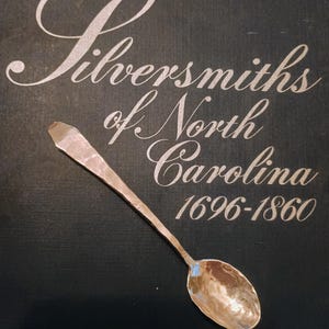 May include: A handcrafted silver spoon with a hammered texture, resting on a dark background. The text "Silversmiths of North Carolina 1696-1860" is elegantly scripted above the spoon, highlighting its historical context.