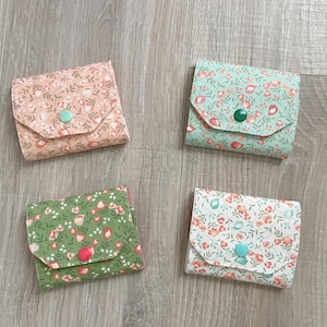 May include: Four small fabric wallets with floral patterns in shades of pink, mint green, dark green, and white. Each rectangular wallet features a triangular flap secured with a single round plastic snap button in a contrasting color.