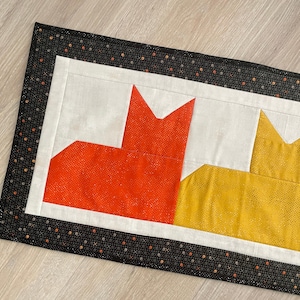 May include: A rectangular quilted table runner featuring three stylized cat silhouettes in orange, yellow, and light green fabric. The cats are set against a white background, framed by a black border patterned with small multicolored dots.