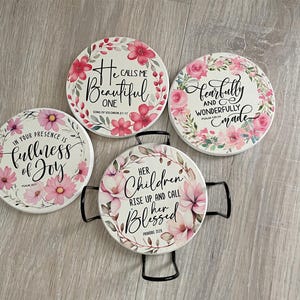 May include: Set of four ceramic coasters with floral designs and inspirational quotes. Each coaster features a different quote in elegant script, surrounded by pink and white flowers. Includes a black metal holder for one coaster.
