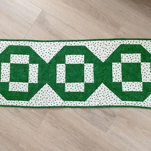 May include: A rectangular green and white quilted table runner. The runner features three large green geometric shapes with white square centers. The white sections are patterned with tiny green hearts. The edges are finished with a green border.