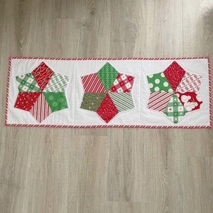 May include: A Christmas-themed table runner featuring three white star-shaped panels. Each star is pieced with red and green patterned fabrics, including stripes, polka dots, and holiday motifs. The runner is bordered with a red and white candy cane stripe.