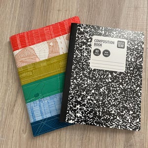 May include: A composition book with a black and white speckled cover and a fabric notebook cover featuring horizontal stripes in red, cream, olive green, bright green, light blue, and dark blue with white stitched line patterns throughout.