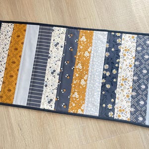 May include: A rectangular quilted table runner featuring a vertical striped pattern of various fabrics in shades of mustard yellow, navy blue, light blue, and white. The design includes floral prints, geometric bee motifs, and thin stripes.