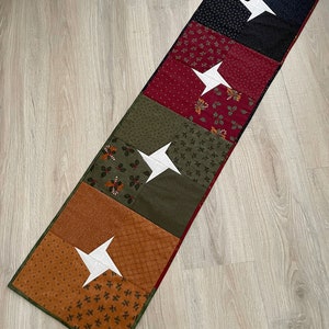 May include: A rectangular table runner with a patchwork design. The runner features alternating squares of various patterns and colors, including black, red, green, and brown. Each square has a white star. The runner is trimmed with a green border.
