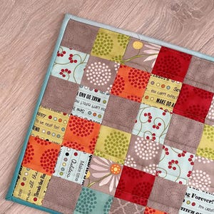 May include: A patchwork quilt with a teal border. The quilt is made of various fabrics with different patterns, including polka dots, flowers, and text. The text on the quilt includes "Make do and", "Sew happy", "Make time for all the little things", and "Love forever!"