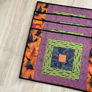May include: A set of four square fabric placemats featuring a layered patchwork design. The patterns include orange pumpkins, black crows, purple spiderwebs, lime green bats, and a dark blue spiderweb center with a solid orange square in the middle.