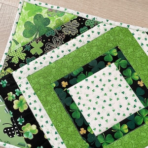May include: A quilted placemat featuring a patchwork design with various green clover patterns on a white and black background. The placemat has a white border with small green clover accents.