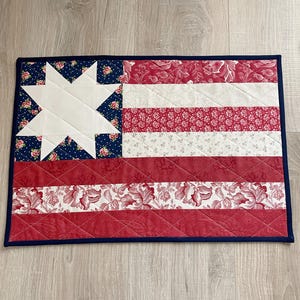 May include: A quilted flag-themed table runner. The design features a white star on a blue floral background, red and white stripes, and a navy blue border. The fabric has a floral pattern.