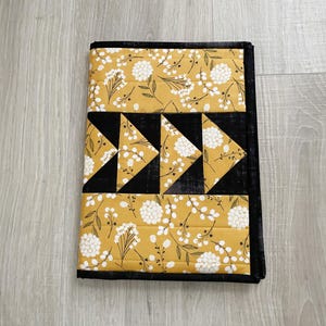 May include: A quilted notebook cover featuring a mustard yellow floral pattern with white flowers and black accents. The design includes black triangles and borders, creating a geometric and botanical aesthetic.