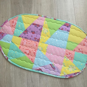 Puede incluir: Una alfombra ovalada de patchwork acolchado con una variedad de telas coloridas. El diseño presenta triángulos y secciones curvas en tonos de rosa, amarillo, verde, azul y morado, con estampados florales y de lunares. La alfombra tiene un borde verde.