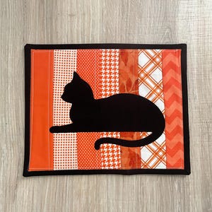 May include: A rectangular fabric mat featuring a black cat silhouette appliqued over vertical strips of orange and white fabric. The background strips display various patterns including polka dots, houndstooth, plaid, and chevron, all framed by a black border.