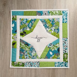 May include: A square, quilted wall hanging with a white center featuring the words "choose JOY" in script. The design incorporates teal, green, and white floral and geometric patterns. The piece is bordered with white fabric.