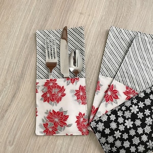 May include: A set of fabric cutlery holders featuring a white pocket with red poinsettia flowers and gray leaves. The upper section has a black and white diagonal striped pattern. One holder contains a stainless steel fork, knife, and spoon.