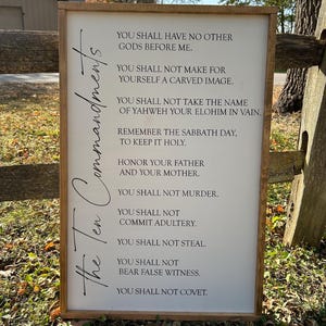 May include: A white wooden sign with a brown frame, featuring the text "The Ten Commandments" written vertically on the left side. The sign lists the Ten Commandments in black text.