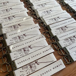 May include: A collection of off-white fabric keychains with bronze-colored hardware. Each keychain is printed with the words "SANDALS CHURCH WOMEN" and a dandelion graphic in a dark brown color. The keychains are arranged on a wooden surface.