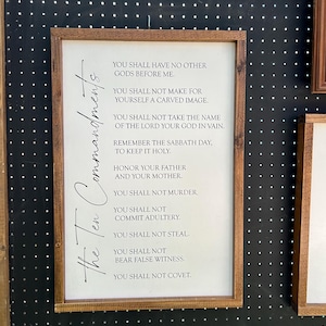May include: A framed wooden sign displaying the Ten Commandments. The text is in black against a white background, with a dark brown wooden frame. The sign is hanging on a black surface with white dots.