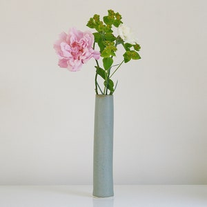 May include: A light grey ceramic vase with a cylindrical shape holds a bouquet of pink and white carnations and green foliage.