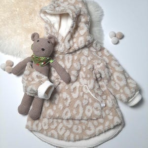 Puede incluir: Una sudadera con capucha beige con estampado de leopardo. La sudadera tiene un bolsillo blanco en la parte delantera y un ribete blanco alrededor de la capucha y el dobladillo.