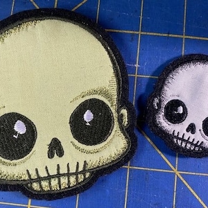 May include: Two embroidered patches featuring cartoon skulls with large eyes. The larger patch is light green with black eyes and a black outline. The smaller patch is light purple with black eyes and a black outline.