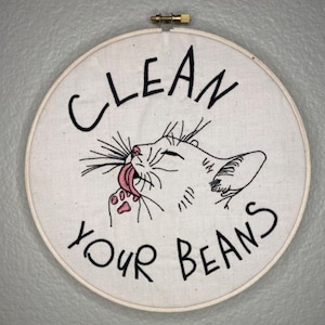 May include: Embroidered hoop art featuring a white cat licking its paw with the text "Clean Your Beans".