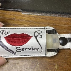 May include: A white and black striped fabric keychain with a clear plastic bottle holder. The fabric has the words "Lip Service" embroidered on it, along with a red lip and a wrench.
