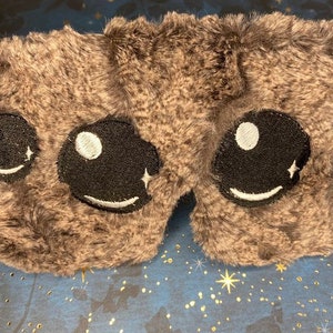 May include: A grey fluffy sleep mask with embroidered black eyes and white stars. The mask is designed to block out light.