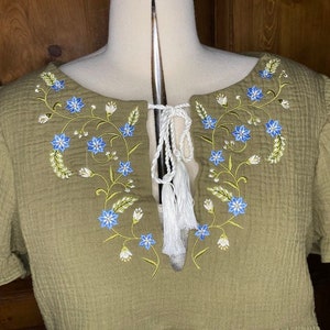 May include: A green cotton top with a V-neck and short sleeves. The top features embroidered floral details in blue and white.