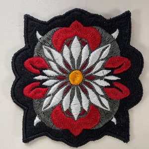 Könnte beinhalten: Ein gestickter Patch in Schwarz, Rot, Weiß und Gelb mit einem Blumendesign. Die Blume hat eine gelbe Mitte und weiße Blütenblätter mit roten Akzenten.