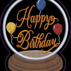 Puede incluir: Diseño bordado de un globo de nieve con el texto "Happy Birthday" en su interior. El globo de nieve tiene una base marrón y tres globos de colores en su interior.