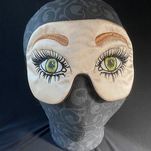 May include: A beige sleep mask with embroidered eyes and eyelashes. The eyes are green and the eyelashes are black. The mask is on a black background.