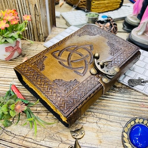 May include: A brown leather journal with a Celtic knot design embossed on the cover. The journal has a metal clasp closure and decorative border. The pages are visible with a yellow edge. The journal is on a wooden surface.