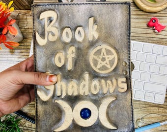 Puede incluir: Un diario de cuero marrón con las palabras "Book of Shadows" grabadas en la portada. La portada también presenta un pentagrama, una luna creciente y una piedra preciosa azul.