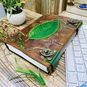 May include: A handcrafted leather journal featuring a green leaf motif on the cover. The journal has a brown cover with green detailing, metal clasps, and a green spine. The pages are white and the journal is closed.