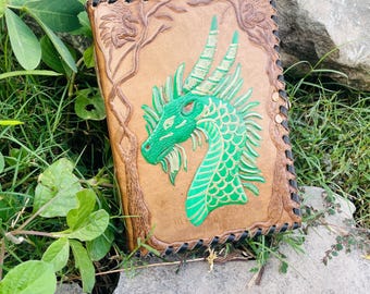 May include: A handmade leather journal with a green dragon design. The dragon is embossed on the front cover, with intricate details and a vibrant green color. The journal has a brown leather cover with a stitched border.