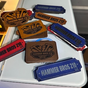 Puede incluir: Un conjunto de parches y etiquetas de cuero en varios colores, incluyendo marrón, rojo y azul. Cada artículo presenta el texto "HAMMER BROS 210" y "HANDYMAN SERVICES" o "LLC", junto con un logotipo de martillo.