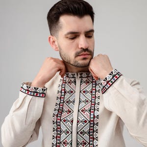 May include: A beige long-sleeved shirt with intricate embroidered patterns in red, black, green, and blue. The embroidery is featured on the collar, cuffs, and down the front of the shirt. The shirt has a high collar.