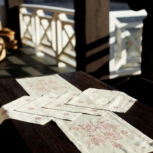 May include: A set of six white linen placemats with embroidered red and gold designs. The placemats are arranged on a dark wooden table.