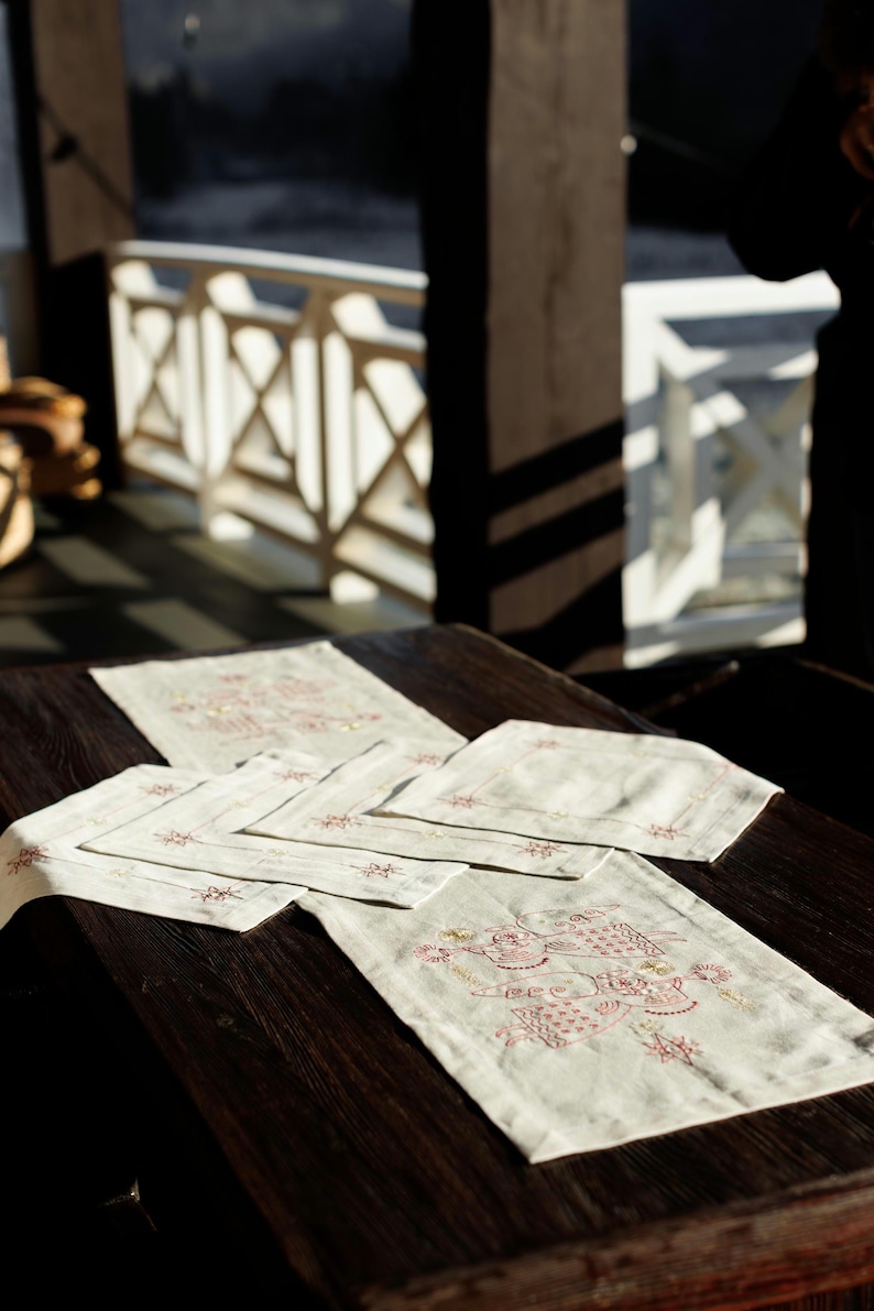 May include: A set of six white linen placemats with embroidered red and gold designs. The placemats are arranged on a dark wooden table.