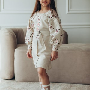 May include: A young girl wearing a beige linen dress with red and green embroidered details. The dress has a belt and long sleeves. She is wearing white socks with bunny ears and black shoes with green bows.