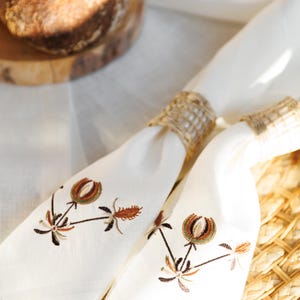 May include: Two white linen napkins with brown and green embroidered floral designs. The napkins are tied together with a brown woven ring.