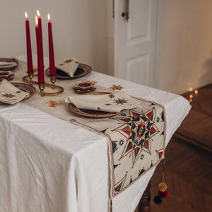 May include: A festive table setting with a white tablecloth, adorned with a star-patterned runner. Red candles in brass holders, plates, and napkins complete the holiday decor. The runner has a star design with red, green, and blue accents.